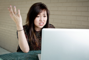 A person appears exasperated as they throw their hand up and look in disbelief at their laptop.