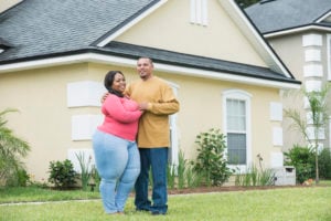 Two people holding one another pose on a front yard in front of a yellow house.