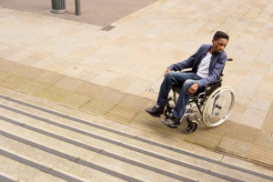 A person in a wheelchair sits sadly at the bottom of some stairs.
