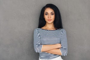 Young adult looking at camera and keeping arms crossed while standing against grey background.