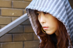 A person sits by a brick wall, looking sad and wearing their hood up.
