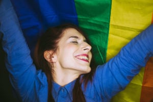 A person smiling with their eyes closed as they hold up a rainbow flag behind them.