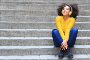 A person sitting on stone stairs, appearing happy as they listen to music from their bright yellow headphones.