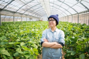 A farmer stands confidently inside a greenhouse, arms crossed and expression certain.