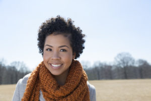 A person wearing a knit scarf stands in a sunny field and smiles at the viewer.