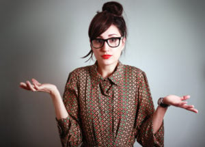 A person with dark-rimmed glasses, bright red lipstick, and a patterned shirt shrugs, looking bewildered.