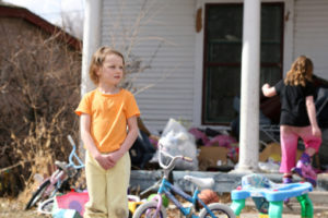 Young child standing in front of a tattered house with bags and other objects scattered in the front yard. Another young child is walking into the house.