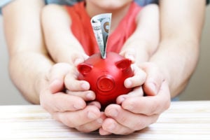 Closeup on a parent and child's hands holding a red piggy bank with a $20 bill sticking out of the top.