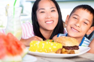 A parent and child smile with a burger, corn on the cob, watermelon, and a glass of lemonade in the foreground.