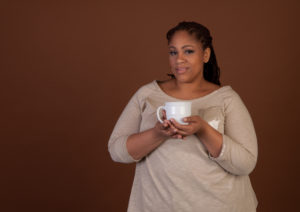 A fat person stands in front of a brown background, holding a cup of coffee and looking perturbed.