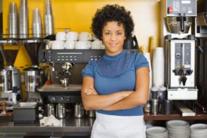 A barista smiles in front of an espresso machine, arms folded.