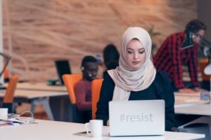 A person wearing a hijab sits at their desk with coworkers in the background. Their laptop reads "#resist." Image source: Thinkstock; text added by Everyday Feminism.