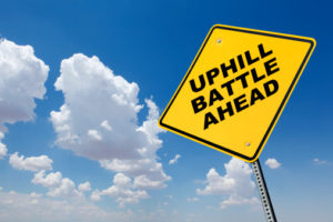 Sign labeled "Uphill Battle Ahead" surrounded by clouds in the sky