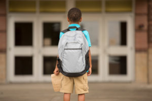 An image of young person from behind with a backpack on while holding a lunch bag 