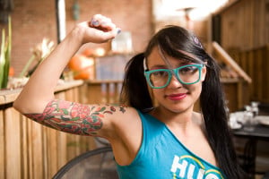 A person with a raised arm with a colorful tattoo on the arm. Their long black hair is in pony-tails.