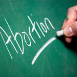 A hand writes the word "Abortion" on a chalkboard.
