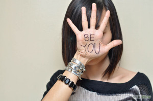 Person with a short, dark bob haircut standing against a wall with their hand in front of their face with "Be You" written on their palm