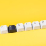 Against a yellow background, keyboard keys spell out "racism." All of the keys are white except for the "A" key, which is black.