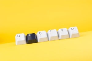 Against a yellow background, keyboard keys spell out "racism." All of the keys are white except for the "A" key, which is black.