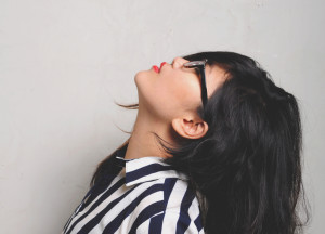 A person wearing glasses, red lipstick, and a striped shirt looking up against a gray background