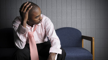 A person sitting in a waiting room has their hand on their head and their tie loosened in frustration and exhaustion