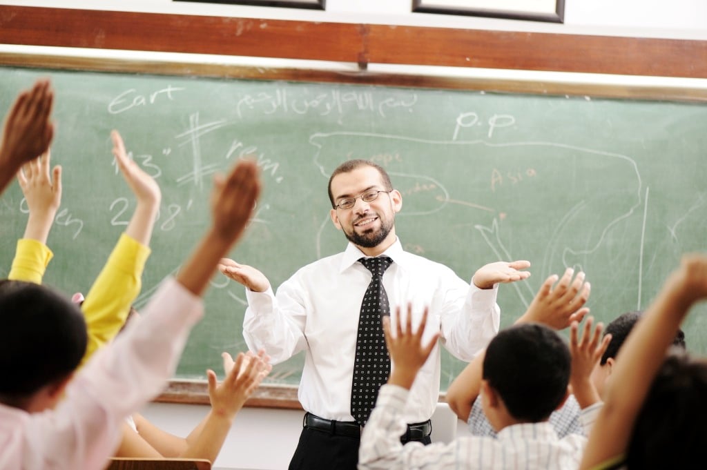 A happy teacher stands at the head of a classroom; many of the childrens' hands are excitedly raised
