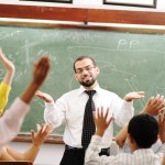 A happy teacher stands at the head of a classroom; many of the childrens' hands are excitedly raised