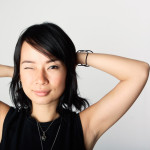 Person looking into camera and winking, with their arms bent up and their hands behind their head, against a gray wall