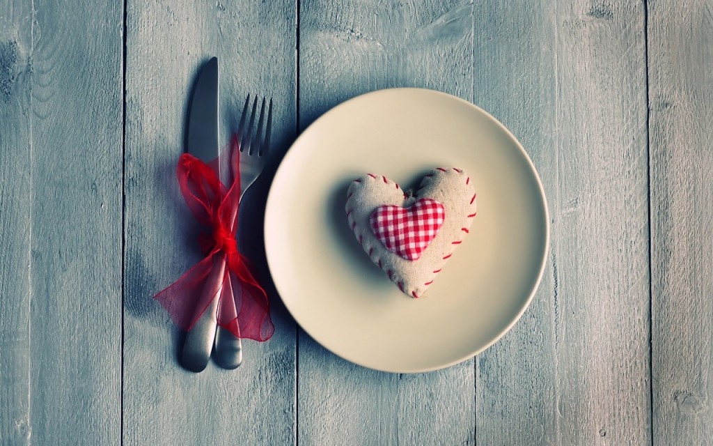 A dinner set-up: A white plate with a heart-shaped pillow on it, a fork and knife tied up with a red ribbon, against a wooden table