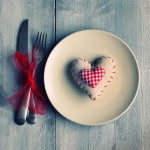 A dinner set-up: A white plate with a heart-shaped pillow on it, a fork and knife tied up with a red ribbon, against a wooden table