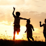 A group of children are silhouetted, playing a ball game against a setting sun