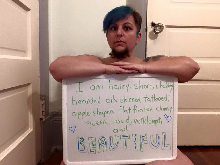 The author sits with a sign. The sign reads, "I am hairy, short, chubby, bearded, oily skinned, tattooed, apple shaped, flat footed, clumsy, queer, loud, verklempt, and BEAUTIFUL."