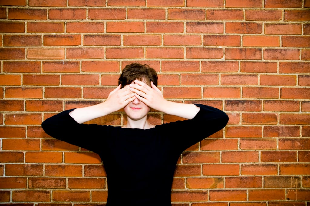 A person standing against a brick wall has their hands over their eyes, covering them