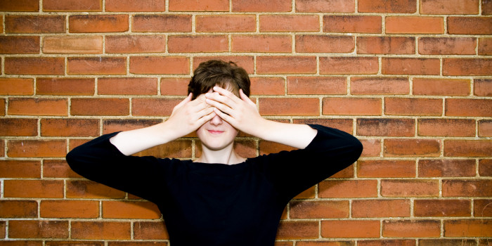 A person standing against a brick wall has their hands over their eyes, covering them