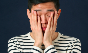 Person in a black-and-white striped shirt against a dark blue background, holding their hands up to their face is disappointment