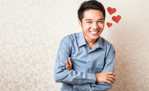 A person in a blue button-up shirt is standing against beige wallpaper, hugging themselves. Hearts are next to their head.