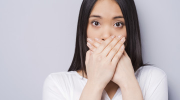A person against a gray background stands with their hands covering their mouth, as if saying "Oops"