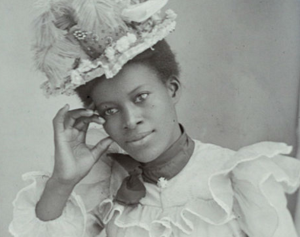 A person in an ornate hat and dress looks pensively into the camera, their hand touching their face.