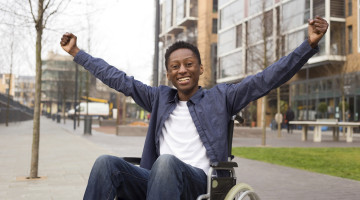A person sits in a wheelchair, stretching out their arms and smiling.