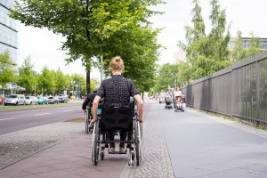 A person in a wheelchair travels down a city sidewalk lined with bright green trees.