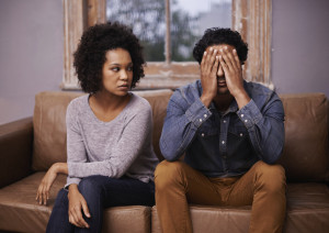 Two people sit beside each other on a couch. One stares at the other, appearing upset. The other has their hands covering their face in distress.