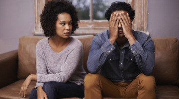 Two people sit beside each other on a couch. One stares at the other, appearing upset. The other has their hands covering their face in distress.