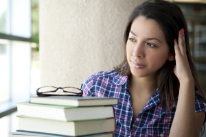 A student sits by a window, next to a stack of books with a pair of glasses on top.
