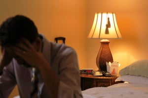 A person sits on the foot of a bed in a hotel room. They are out of focus; the camera focuses in on a bedside table, which has a lamp, alarm clock, and takeout container.