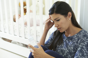 A person is sitting, looking distressed with their hand touching their forehead, scrutinizing a formula bottle. A crib is behind them with an infant sleeping.