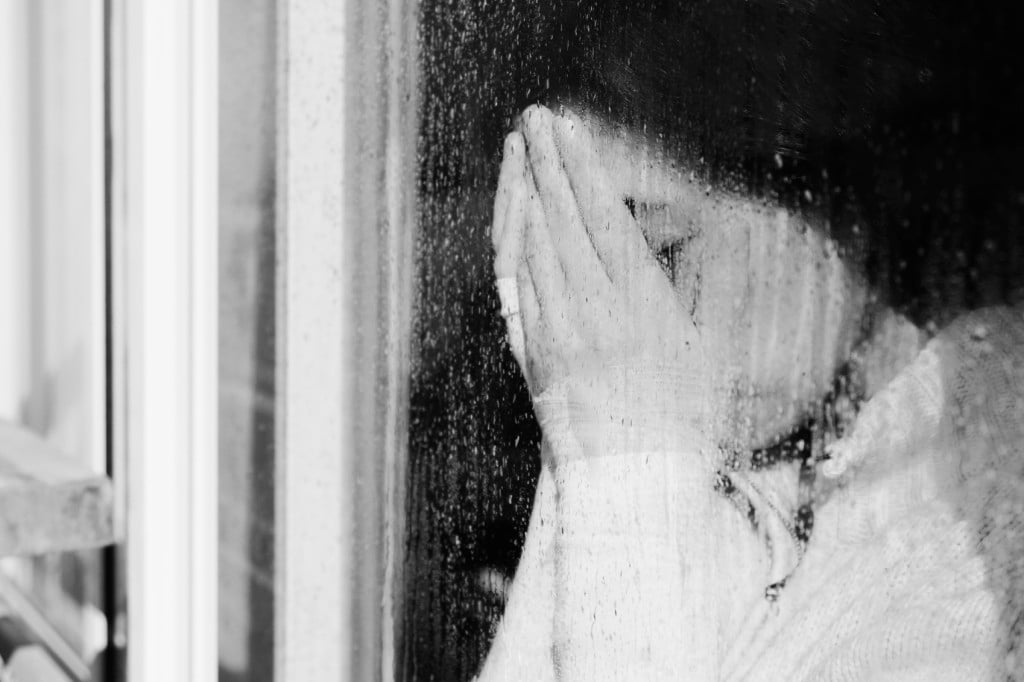 A person is covering their face as they stand beside a window covered in raindrops.