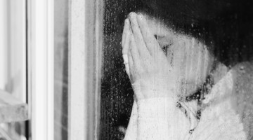 A person is covering their face as they stand beside a window covered in raindrops.