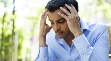 A stressed person has their hands on head, sitting outside.