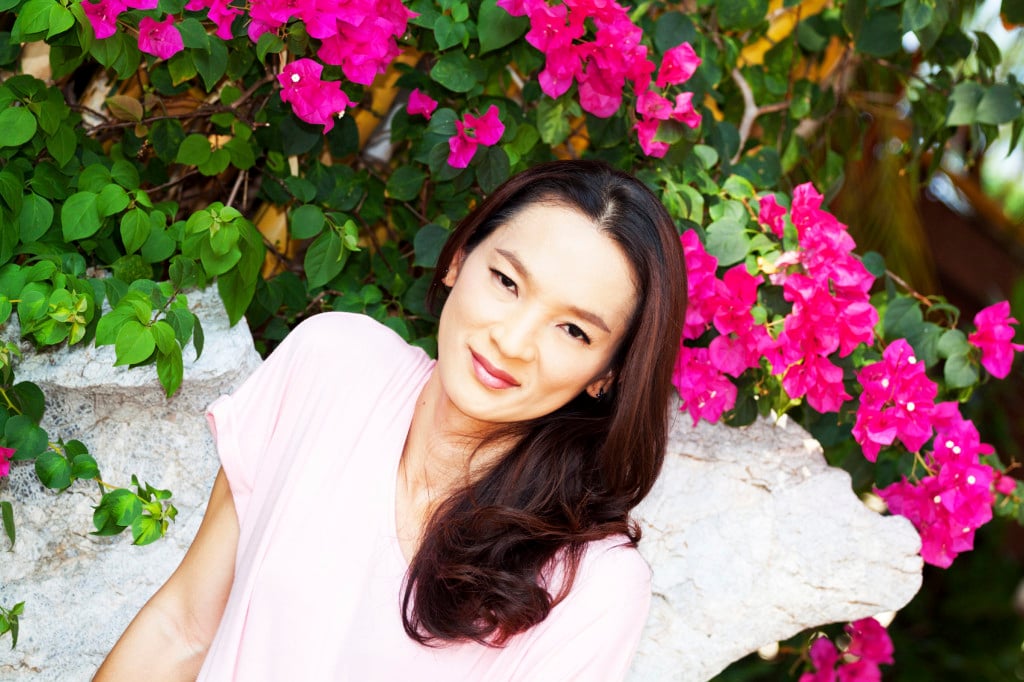 A person sits, smiling, in front of a wall of bright pink flowers.