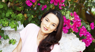 A person sits, smiling, in front of a wall of bright pink flowers.